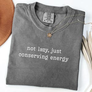 Può includere: T-shirt grigio Comfort Colors con la scritta bianca "not lazy, just conserving energy". Una collana color oro con un piccolo ciondolo è drappeggiata sulla maglietta. Un cappello marrone è visibile in alto a sinistra.