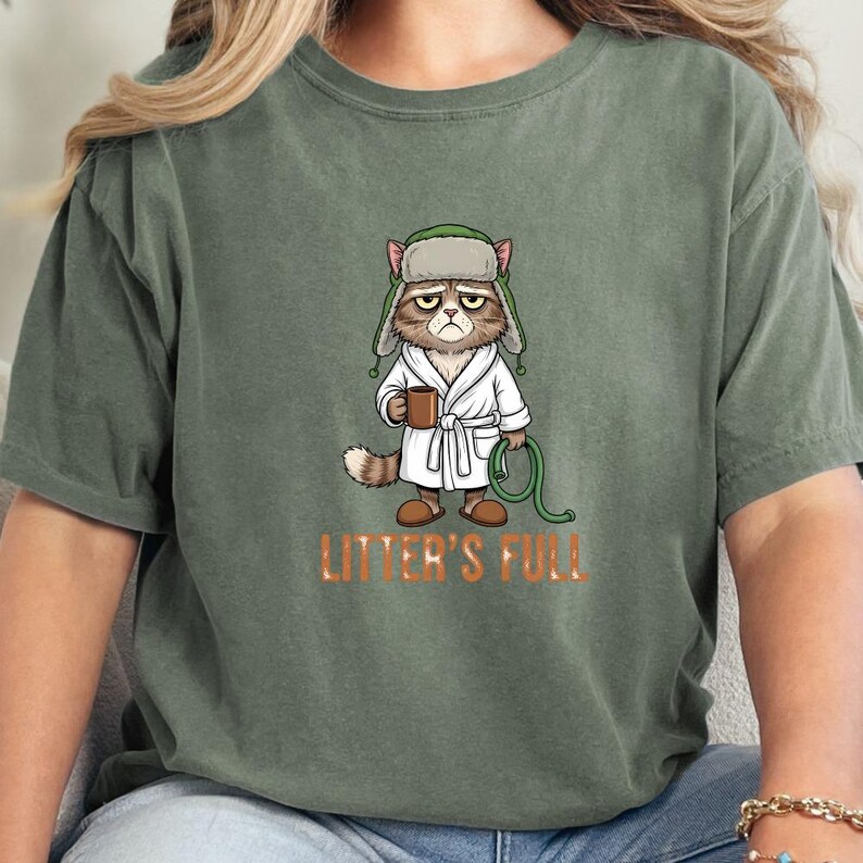 May include: Olive green t-shirt featuring a grumpy cat cartoon wearing a hat, robe, and holding a mug and a green hose. The text "LITTER'S FULL" is printed in orange below the cat. The cat has a disgruntled expression.