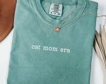 Könnte beinhalten: Ein meergrünes T-Shirt mit dem weißen Schriftzug "cat mom era". Eine goldfarbene Halskette mit einem kleinen Anhänger liegt auf dem Shirt. Das Shirt hat einen Rundhalsausschnitt und ein Comfort Colors-Etikett.