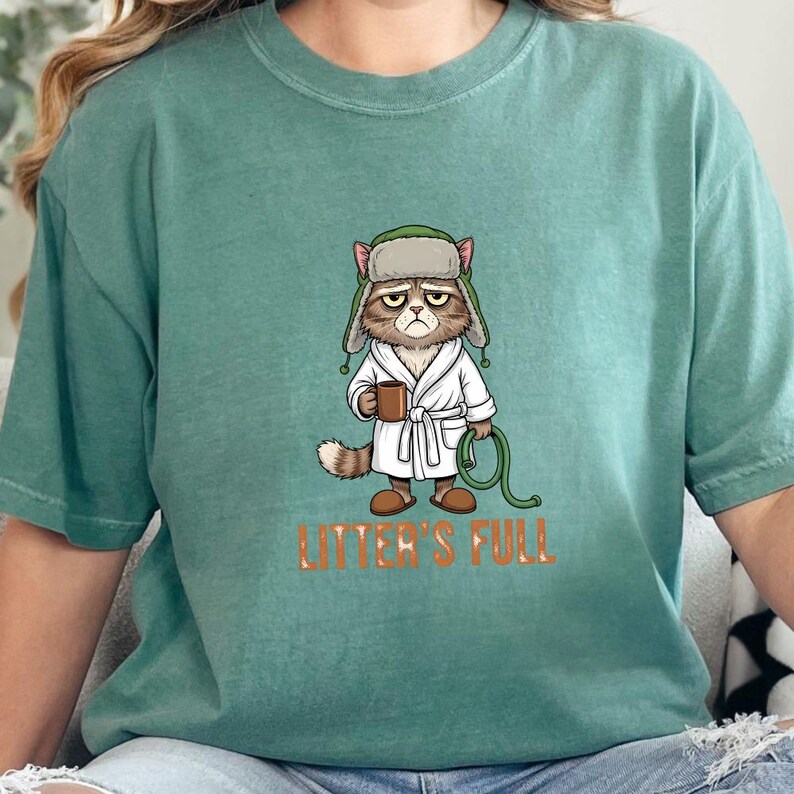 May include: A seafoam green t-shirt featuring a grumpy cat cartoon wearing a hat and robe, holding a mug and a hose. The text "LITTER'S FULL" is printed in orange below the cat illustration.