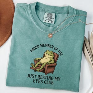 May include: A teal Comfort Colors t-shirt with a graphic of a frog relaxing in a chair. The text reads "Proud Member of the Just Resting My Eyes Club." A gold necklace with a pendant rests on the shirt.
