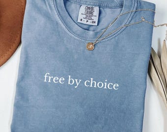Może przedstawiać: Niebieska koszulka Comfort Colors z białym napisem "free by choice". Złoty naszyjnik z wisiorkiem spoczywa na koszulce. Koszulka jest starannie złożona.
