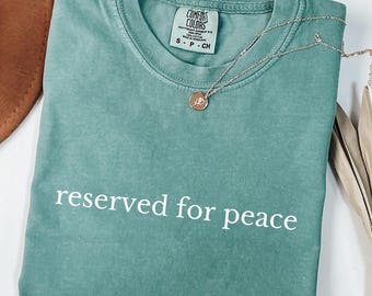 Może przedstawiać: Zielona koszulka Comfort Colors z napisem "reserved for peace" w białych literach. Złoty naszyjnik z małą zawieszką spoczywa na koszulce. Koszulka jest starannie złożona.