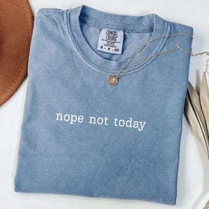 Könnte beinhalten: Blaues Comfort Colors T-Shirt mit dem weißen Schriftzug „nope not today“. Eine goldene Halskette mit Anhänger liegt auf dem gefalteten Shirt. Das Shirt hat ein Etikett.