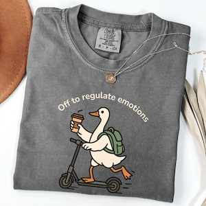 Puede incluir: Una camiseta gris Comfort Colors con un pato de dibujos animados en un scooter, sosteniendo una taza de café. El texto "Off to regulate emotions" está encima del pato. Un collar plateado con un colgante está sobre la camiseta.