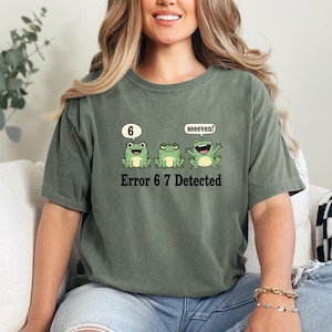 May include: Olive green t-shirt featuring three cartoon frogs. The first frog has a speech bubble with the number 6, the second frog is neutral, and the third frog has a speech bubble that says "seeeven!". The text "Error 6 7 Detected" is below the frogs.