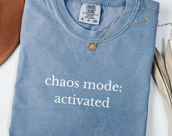 May include: A blue Comfort Colors t-shirt with the text "chaos mode: activated" in white. A gold necklace with a pendant is draped over the folded shirt. The shirt has a small tag at the neckline.