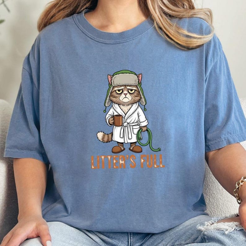 May include: A light blue t-shirt featuring a grumpy cat cartoon wearing a robe and hat, holding a mug and a green hose. The text "LITTER'S FULL" is printed in orange below the cat.