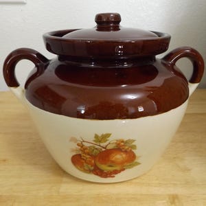 May include: A brown and cream-colored ceramic crock pot with a lid and two handles. The crock pot has a fruit and leaf design on the cream-colored base. The lid and top portion of the crock pot are brown.