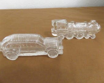 May include: Two clear glass figurines shaped like vehicles. One figurine is a vintage-style automobile with a ribbed roof and defined wheels. The second figurine is a steam locomotive train with a prominent boiler, cab, and multiple wheel details.