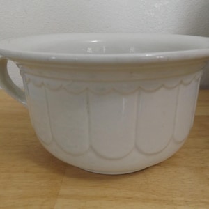 May include: A white ceramic chamber pot featuring a wide, flared rim and a single side handle. The exterior body is decorated with a repeating scalloped pattern that extends from the base up to the smooth, rounded upper section of the vessel.