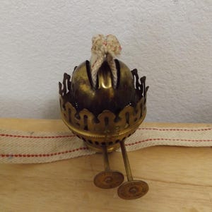 May include: A brass oil lamp burner featuring a decorative scalloped gallery, a central wick holder with a white cotton wick, and two small adjustment knobs extending from the base, accompanied by a flat white fabric wick with red stitching.