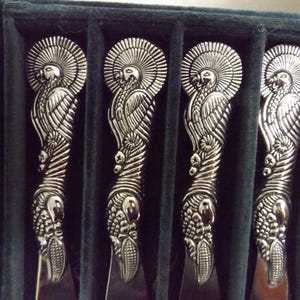 May include: Set of four silver-toned knives with ornate handles featuring a bird design. The knives are displayed in a dark green velvet-lined case, showcasing the intricate details of the cutlery.