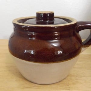 May include: A two-tone ceramic crock with a lid and handle. The upper portion is a glossy, dark brown, while the lower half is a creamy beige. The lid has a small knob. This vintage-style crock is suitable for kitchen storage.