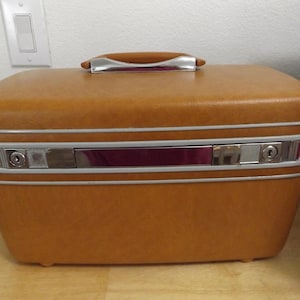 May include: A rectangular, mustard-colored hard-shell vanity case with a textured finish. It features a top handle with chrome accents, a central chrome-colored locking mechanism with two keyholes, and light gray trim bordering the middle section.
