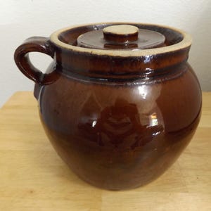 Puede incluir: Un tarro de cerámica marrón con tapa y asa. El tarro tiene un acabado brillante y una forma redondeada. La tapa es circular con un pequeño pomo. El asa es un lazo curvado. Para el almacenamiento de alimentos.