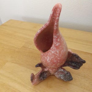 May include: A pink ceramic vase with a speckled pattern and a unique, organic shape. The vase has a wide opening and a dark brown base, resembling a stylized flower or abstract form. The vase is a decorative item.