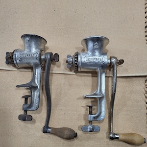 May include: Two vintage Universal meat grinders. The grinders are made of silver-colored metal with a clamp base and a hand crank with a wooden handle. The word "UNIVERSAL" is embossed on the side of each grinder.
