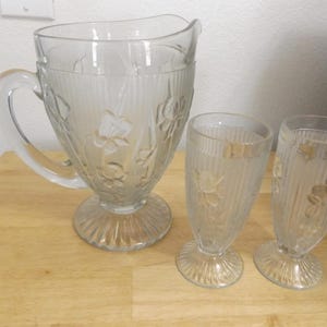 May include: A clear glass pitcher and two matching glasses. The pitcher has a large handle and a wide spout. All pieces feature a floral design and vertical ribbed detailing. The pitcher and glasses sit on a wooden surface.