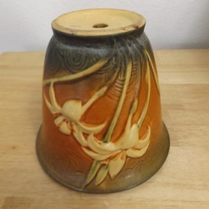 May include: A bell-shaped ceramic lamp shade featuring a relief design of cream-colored flowers and stems against a gradient background that transitions from deep brown at the top to a warm orange hue at the base, with a circular opening at the top.