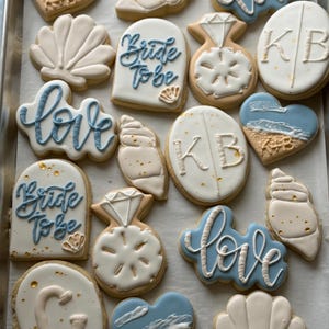Puede incluir: Una bandeja de galletas de azúcar decoradas con temática de boda. Los diseños incluyen conchas marinas, corazones, anillos de compromiso y galletas con las palabras "Bride To Be" y "love" en glaseado azul. Algunas galletas tienen iniciales.