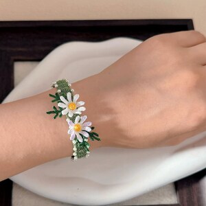 May include: A handmade daisy bracelet featuring white and yellow flower accents with green leaves and a green band. The bracelet is adorned with small white beads and is worn on a wrist.