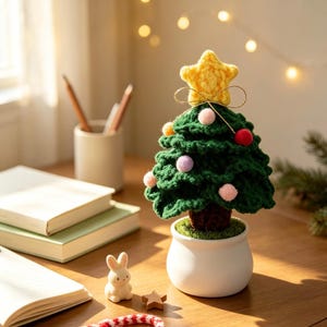 以下が含まれることがあります： 黄色い星のトップが付いたダークグリーンのクロッシェ編みのクリスマスツリー。ピンク、赤、オレンジの小さなポンポンで飾られています。茶色の幹を持ち、白い陶器の鉢に植えられ、本やウサギのフィギュアの近くの木製の表面に置かれています。