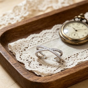 May include: A silver-toned, open-ended ring with a crisscross design, encrusted with small, clear gemstones. The ring rests on a white lace doily inside a wooden tray, with a vintage pocket watch in the background.