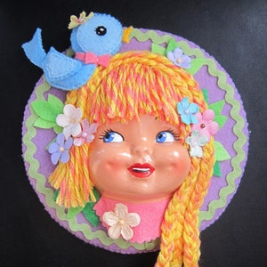 May include: A felt craft depicting a girl with long yellow and pink braids, wearing a purple hat with green leaves and flowers. A blue bird with a bow sits on top of the hat.