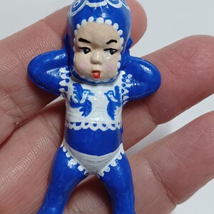 May include: A blue and white figurine of a baby-like figure with arms raised. The figurine has a blue hat and outfit with white trim and accents. The face is a light skin tone with red lips. A silver-colored loop is at the top.
