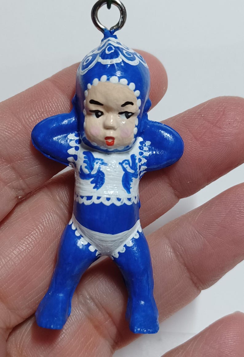 May include: A blue and white figurine of a baby-like figure with arms raised. The figurine has a blue hat and outfit with white trim and accents. The face is a light skin tone with red lips. A silver-colored loop is at the top.