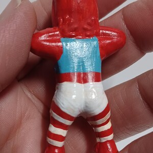 May include: A small, handmade figurine of a person with a red head and arms raised. The figurine wears a blue tank top and white shorts with red stripes. A green string is attached to the top of the head.