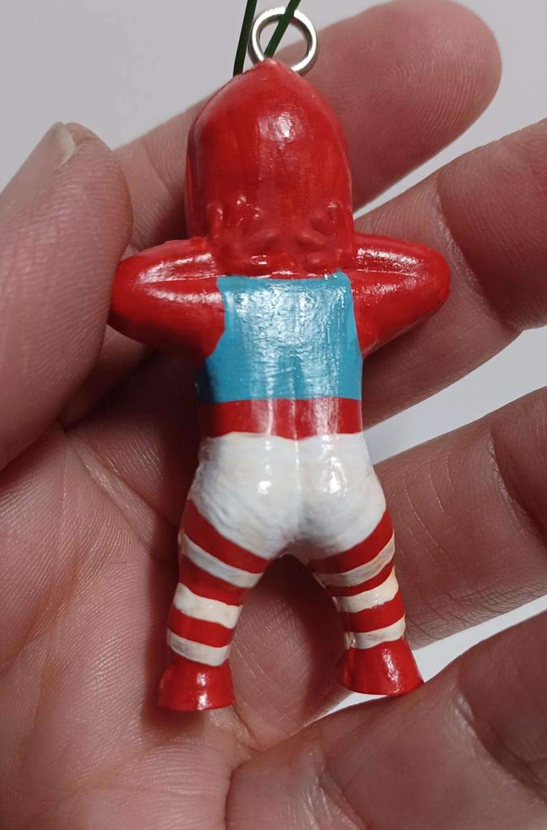 May include: A small, handmade figurine of a person with a red head and arms raised. The figurine wears a blue tank top and white shorts with red stripes. A green string is attached to the top of the head.
