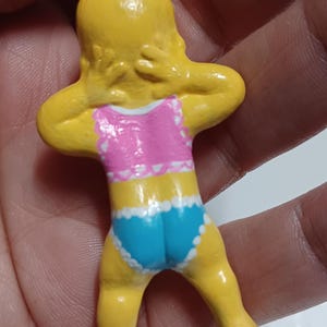 May include: A yellow figurine of a person in a pink and blue swimsuit. The figurine has its arms behind its head. The swimsuit has white trim. The figurine is attached to a silver metal loop.