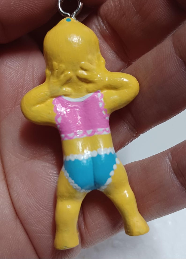 May include: A yellow figurine of a person in a pink and blue swimsuit. The figurine has its arms behind its head. The swimsuit has white trim. The figurine is attached to a silver metal loop.