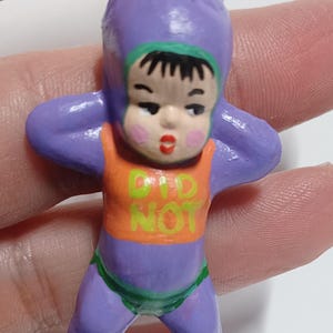 May include: A small, hand-painted figurine of a baby in a purple onesie with an orange top. The text "DID NOT" is printed on the top. The baby has black hair, a green trim, and is holding its arms up. The figurine is attached to a metal loop.
