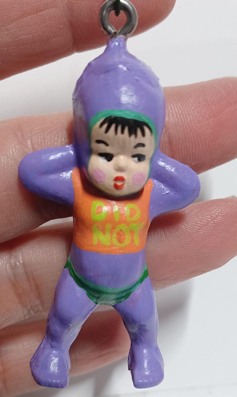 May include: A small, hand-painted figurine of a baby in a purple onesie with an orange top. The text "DID NOT" is printed on the top. The baby has black hair, a green trim, and is holding its arms up. The figurine is attached to a metal loop.