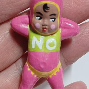 May include: A pink figurine of a baby with a yellow and green top that says "NO". The baby is wearing a pink hood and yellow diaper. The baby's arms are raised to its head.