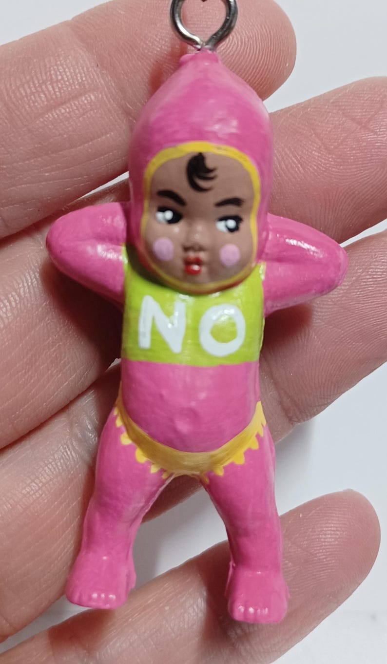 May include: A pink figurine of a baby with a yellow and green top that says "NO". The baby is wearing a pink hood and yellow diaper. The baby's arms are raised to its head.