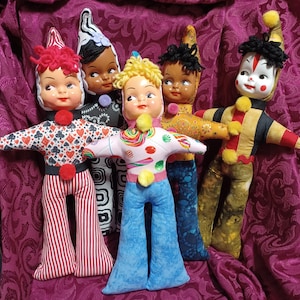 May include: A group of six vintage clown dolls with painted faces, each wearing unique fabric outfits. The dolls have different colored hair and hats. The outfits include patterns like playing cards, stripes, and swirls. Each doll has a pom-pom on the chest and a matching one on the back.