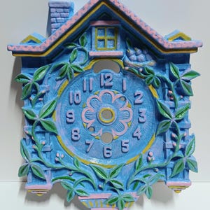 May include: A blue, pink, and yellow decorative cuckoo clock with a floral design. The clock features a house-shaped frame with a chimney, window, and a bird. The clock face has a floral center and the numbers 1 through 12.