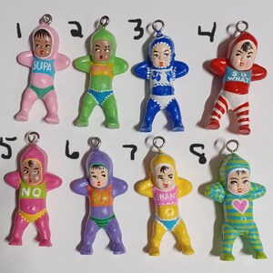 May include: A collection of eight vintage-style baby doll charms. Each doll is painted in a different color, including pink, green, blue, and yellow. The dolls have a hood and are wearing swimsuits. Some dolls have text on their chest.