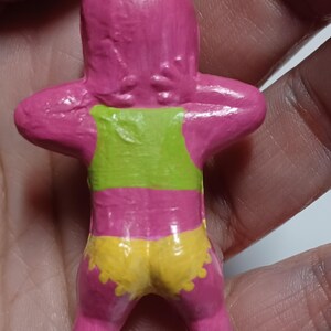 May include: A handmade pink figurine with a green top and yellow shorts. The figurine has its arms raised behind its head. A small metal loop is attached to the top of the figurine.