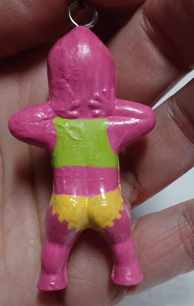 May include: A handmade pink figurine with a green top and yellow shorts. The figurine has its arms raised behind its head. A small metal loop is attached to the top of the figurine.