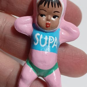 May include: A small, vintage figurine of a baby in a pink onesie with a matching hood. The baby has dark hair and is wearing a blue shirt with the word "SUPA" in white. The baby is also wearing green underwear. The figurine has a silver metal loop at the top.