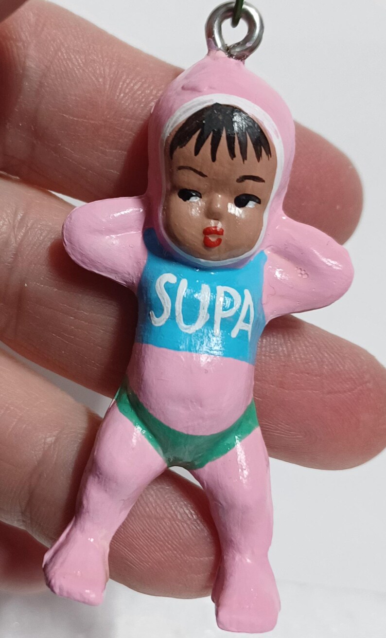 May include: A small, vintage figurine of a baby in a pink onesie with a matching hood. The baby has dark hair and is wearing a blue shirt with the word "SUPA" in white. The baby is also wearing green underwear. The figurine has a silver metal loop at the top.