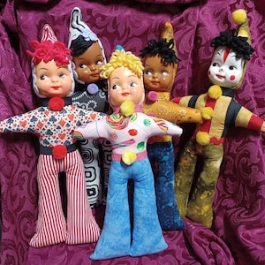 May include: A group of six vintage-style dolls with painted faces and fabric bodies. The dolls wear colorful outfits, including patterned tops and bell-bottom pants. Each doll has a unique hairstyle and hat, with pom-poms and other decorative elements.
