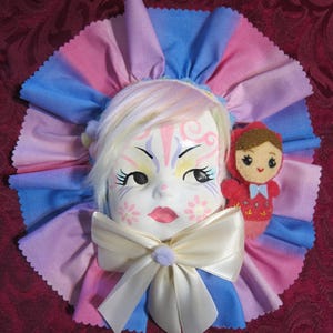 May include: A white porcelain doll face with painted blue and pink details, wearing a white satin bow and a pink and blue fabric ruffle collar. A small red doll with a white and blue dress is attached to the collar.