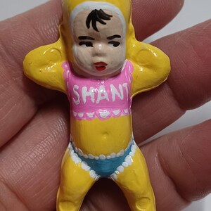 May include: A yellow figurine of a baby with a pink top that says "SHANT" and blue underwear. The baby has black hair and red lips. The figurine is held by a silver loop.