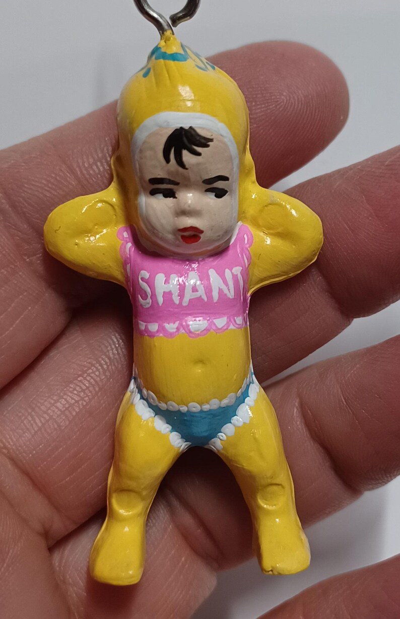 May include: A yellow figurine of a baby with a pink top that says "SHANT" and blue underwear. The baby has black hair and red lips. The figurine is held by a silver loop.